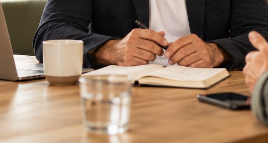 Hände am Tisch mit Notizbuch, Laptop und Wasserglas — ein Gespräch im Betrieb